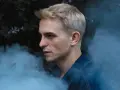 Vito Bambino w dymie, zapowiada nową płytę. Artysta w denimowej kurtce, z blond włosami, na tle ciemnej roślinności.