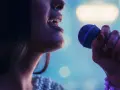 Kobieta śpiewa do mikrofonu. Czy to playback? Wokalistka z błyszczącą suknią i fioletowym mikrofonem.