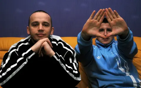 Dwóch mężczyzn w dresach, jeden z rękami złożonymi na brodzie, drugi tworzy rękami kształt serca. Klimat hip hop i rap.