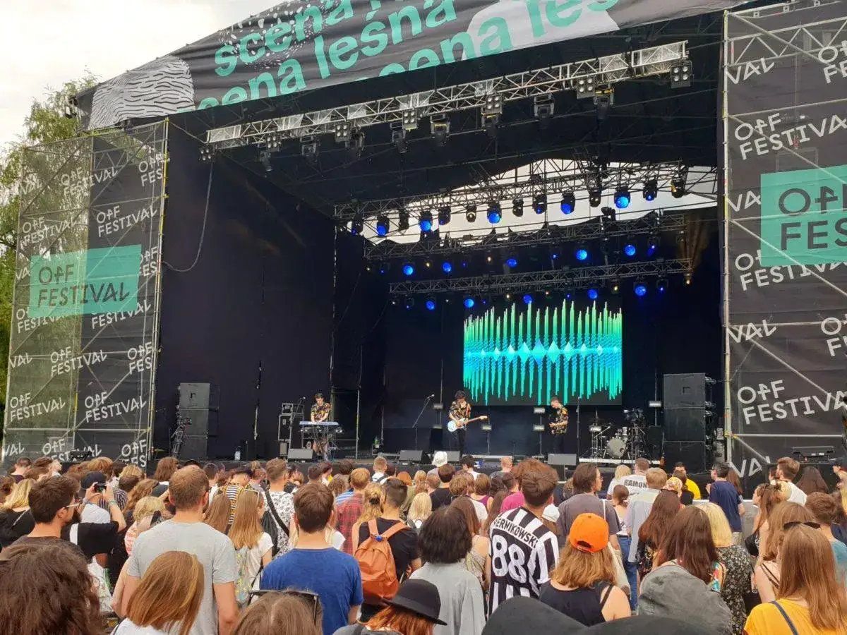 Tłum ludzi na koncercie OFF Festival. Na scenie gra zespół, a za nim wyświetla się kolorowy wizualizer.